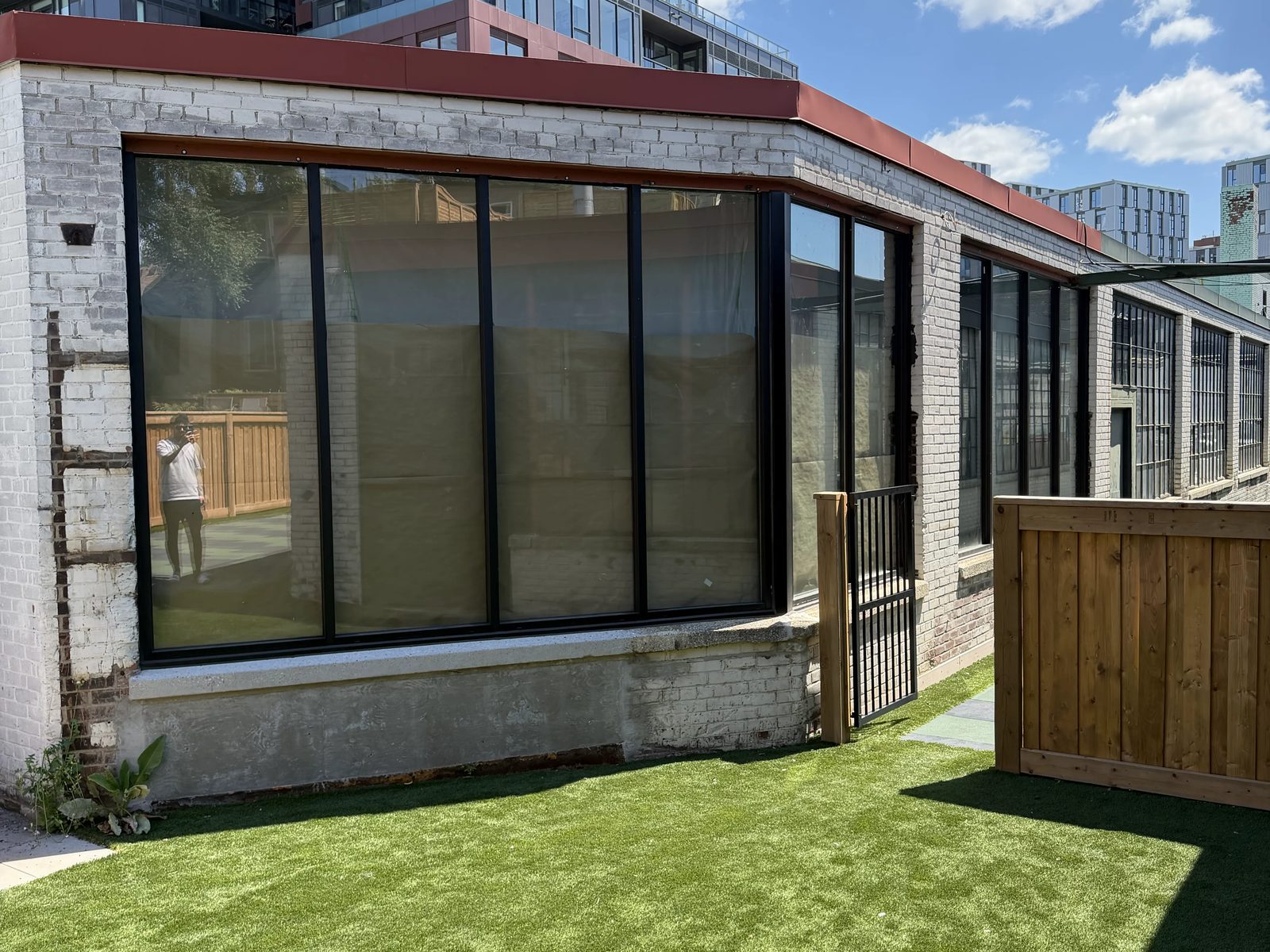 A band of new black-framed aluminum windows wrapping the corner of a whitewashed brick building above a turf play yard in Toronto, installed by Canuck Door Systems