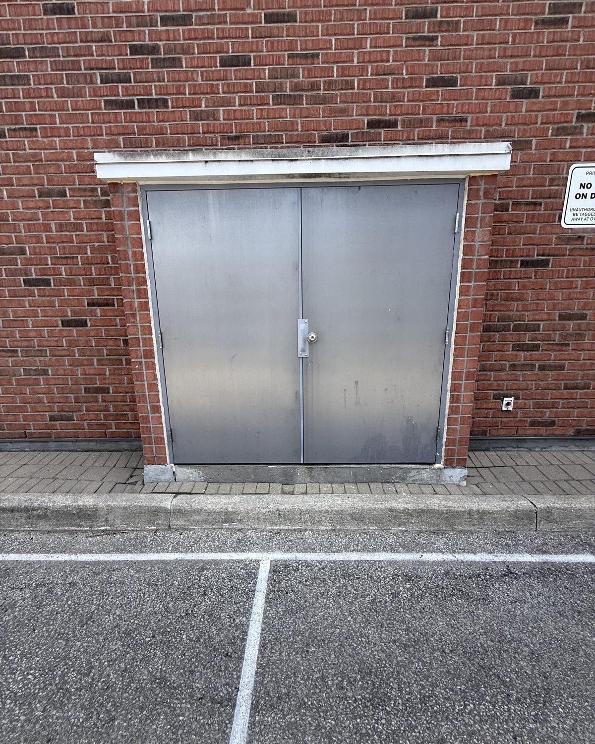 A pair of grey hollow metal doors set into a red brick wall above a paved parking area, with a sign on the wall beside them
