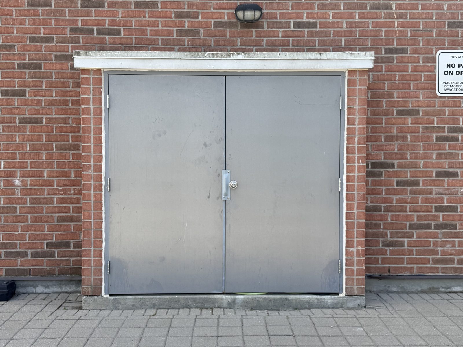 A new pair of grey hollow metal doors under a white head flashing, set into the red brick wall of an electrical room in a Toronto townhome community, installed by Canuck Door Systems