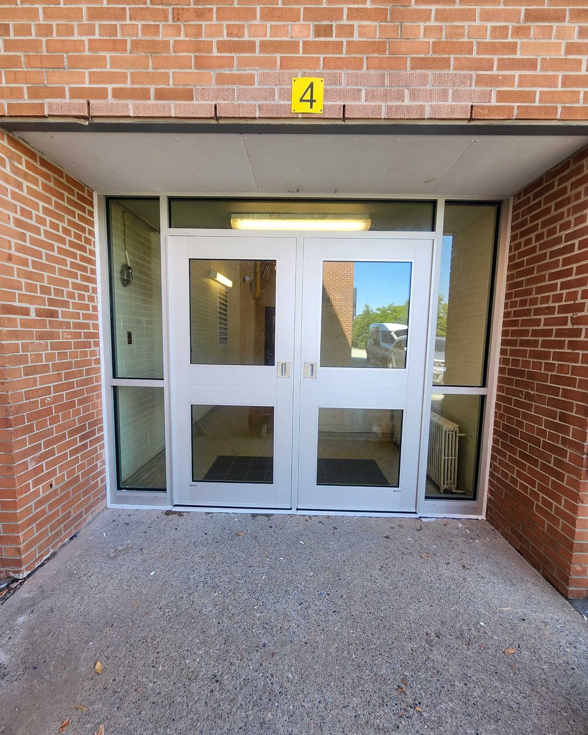 A recessed entrance at St. Albert Catholic School in Toronto: new aluminum double doors with glazed upper and lower lites, glazed sidelites either side and a transom above, under a brick overhang