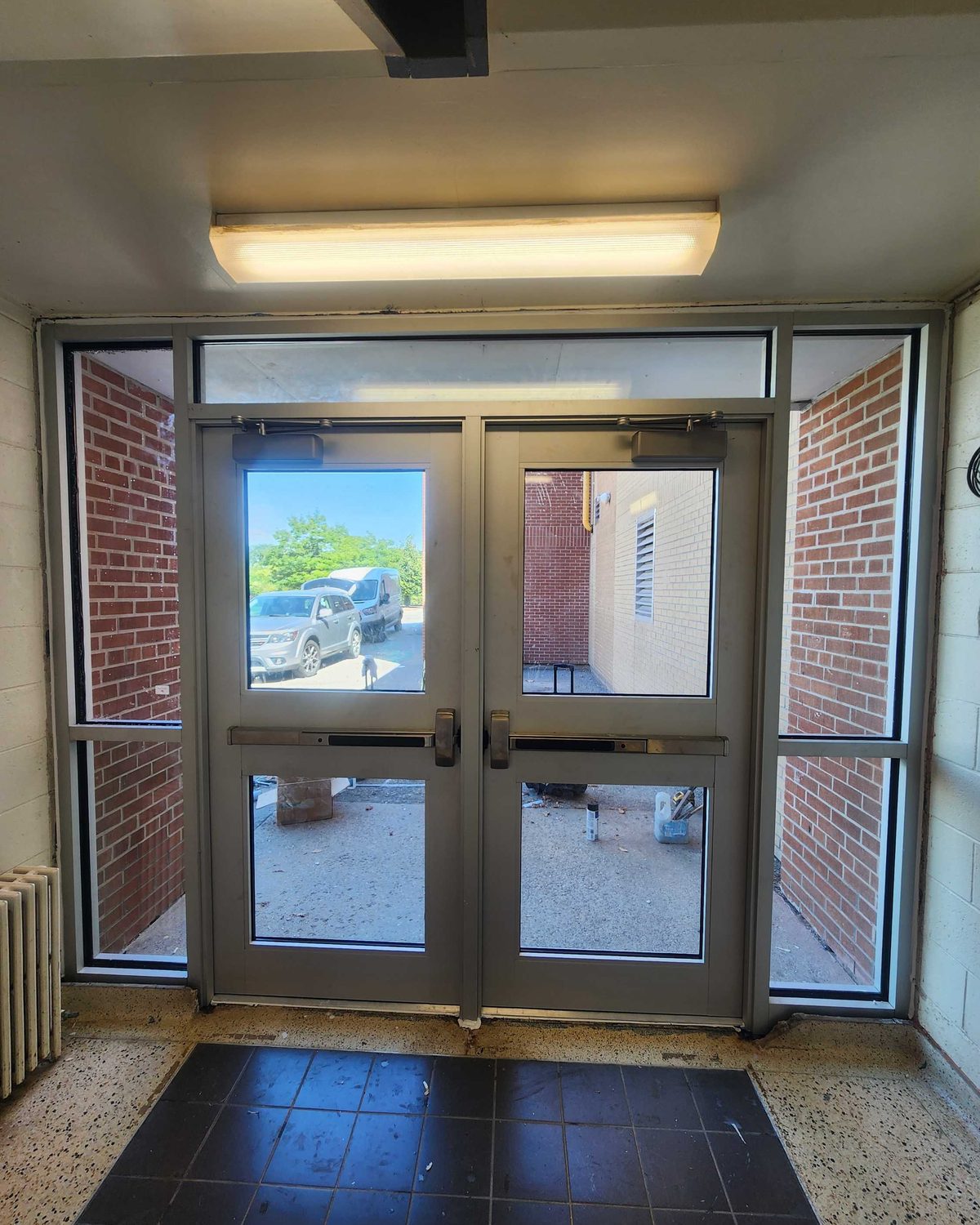 The same kind of aluminum double-door entrance seen from inside a school vestibule, with glazed sidelites and a transom and daylight coming through the glass
