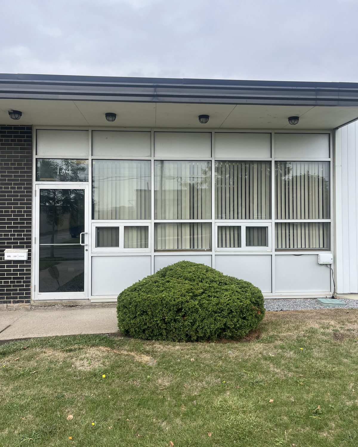 A clear anodized aluminum storefront with a glazed door at the end of a commercial unit on Sunrise Avenue, Toronto, between dark brick and white panels