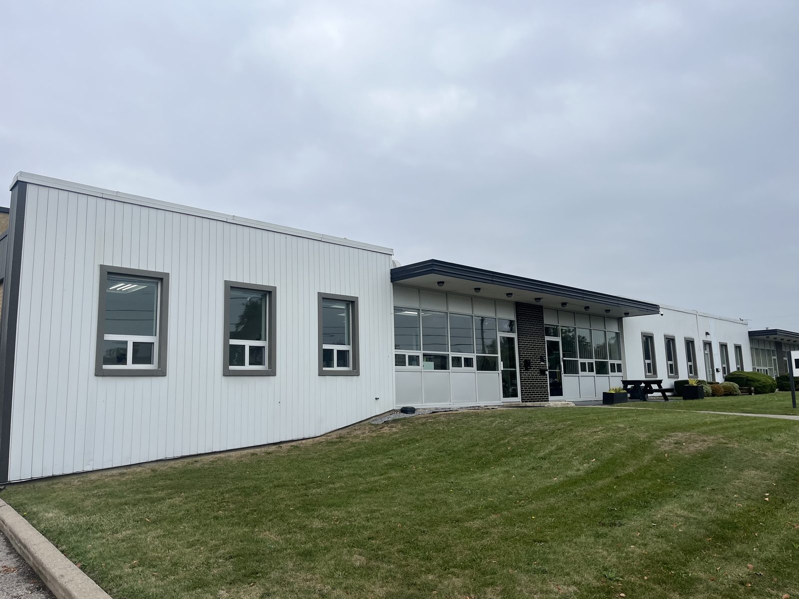 A single-storey white-clad commercial building on Sunrise Avenue in Toronto, with new sliding windows along the side wall and glazed storefronts under the canopy beyond, installed by Canuck Door Systems