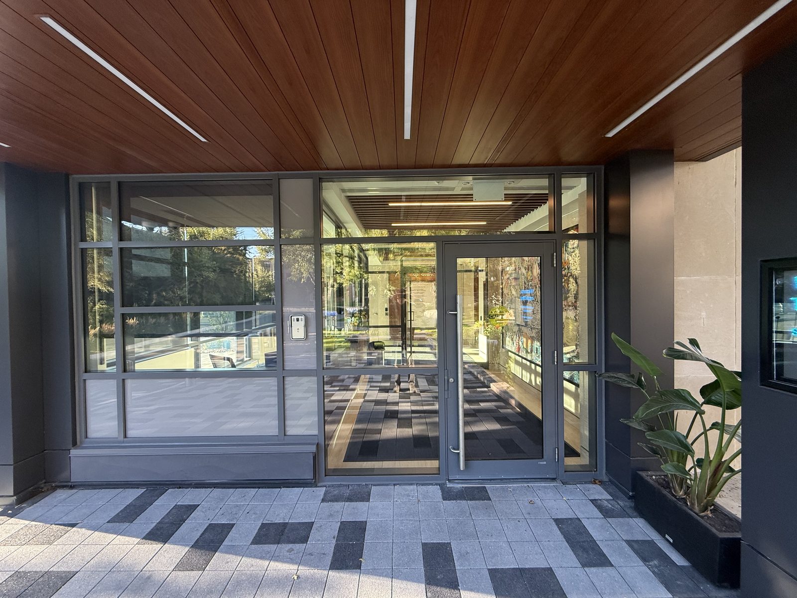 The new west entrance at Temple Sinai Congregation in Toronto: grey aluminum curtain wall glazing and glass doors under a wood-slat canopy, with the vestibule visible behind the glass, installed by Canuck Door Systems