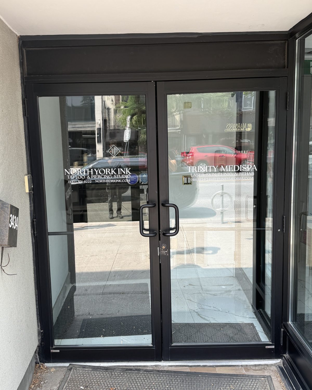 A pair of glass entrance doors in dark aluminum framing, recessed under a canopy at 3434 Yonge Street in Toronto