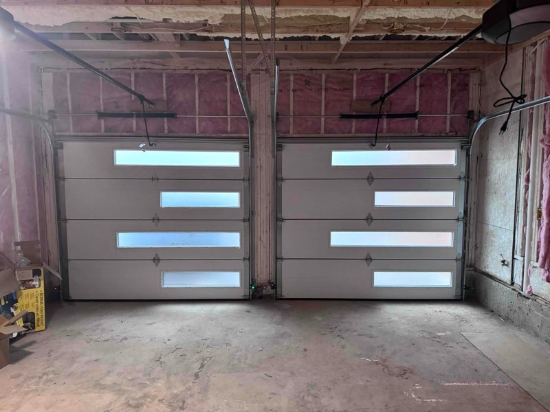 Two beige sectional garage doors with glazed panel slots, newly fitted side by side in an unfinished garage, with the opener rails and the shafts above the door heads visible against the exposed studs and insulation
