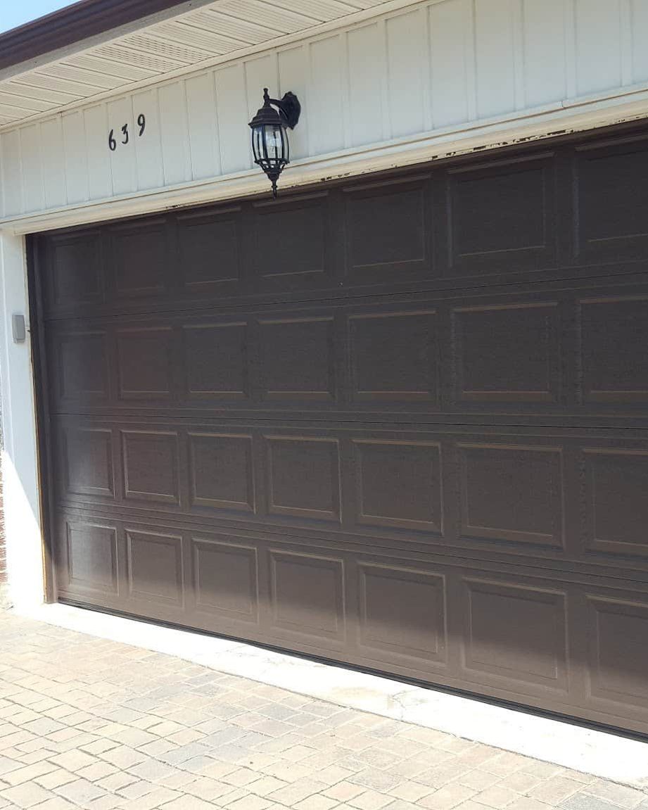 A brown raised-panel steel double garage door closed in its white-trimmed opening on a brick house, with a coach light and the house number above it and a paver driveway in front