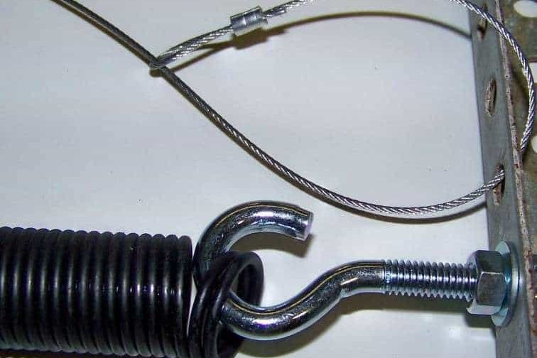 The hooked end of an extension spring anchored to the track bracket, with a steel containment cable threaded through the spring and running past the hook