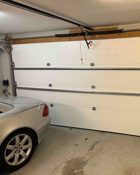 A wide white sectional garage door seen from inside a home garage, with the opener rail, the torsion shaft and the red emergency release cord above it and a car parked alongside