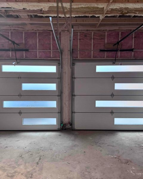 A pair of glazed-panel garage doors seen from inside an unfinished garage, with the track and spring hardware above them and exposed framing overhead