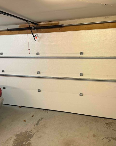 A white insulated double garage door from inside a garage, with the opener rail running back along the ceiling, the torsion shaft and spring above the door head, and a car parked alongside
