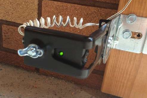 A garage door safety photo eye in its bracket, mounted low on a brick wall beside the door track, with its green indicator light lit