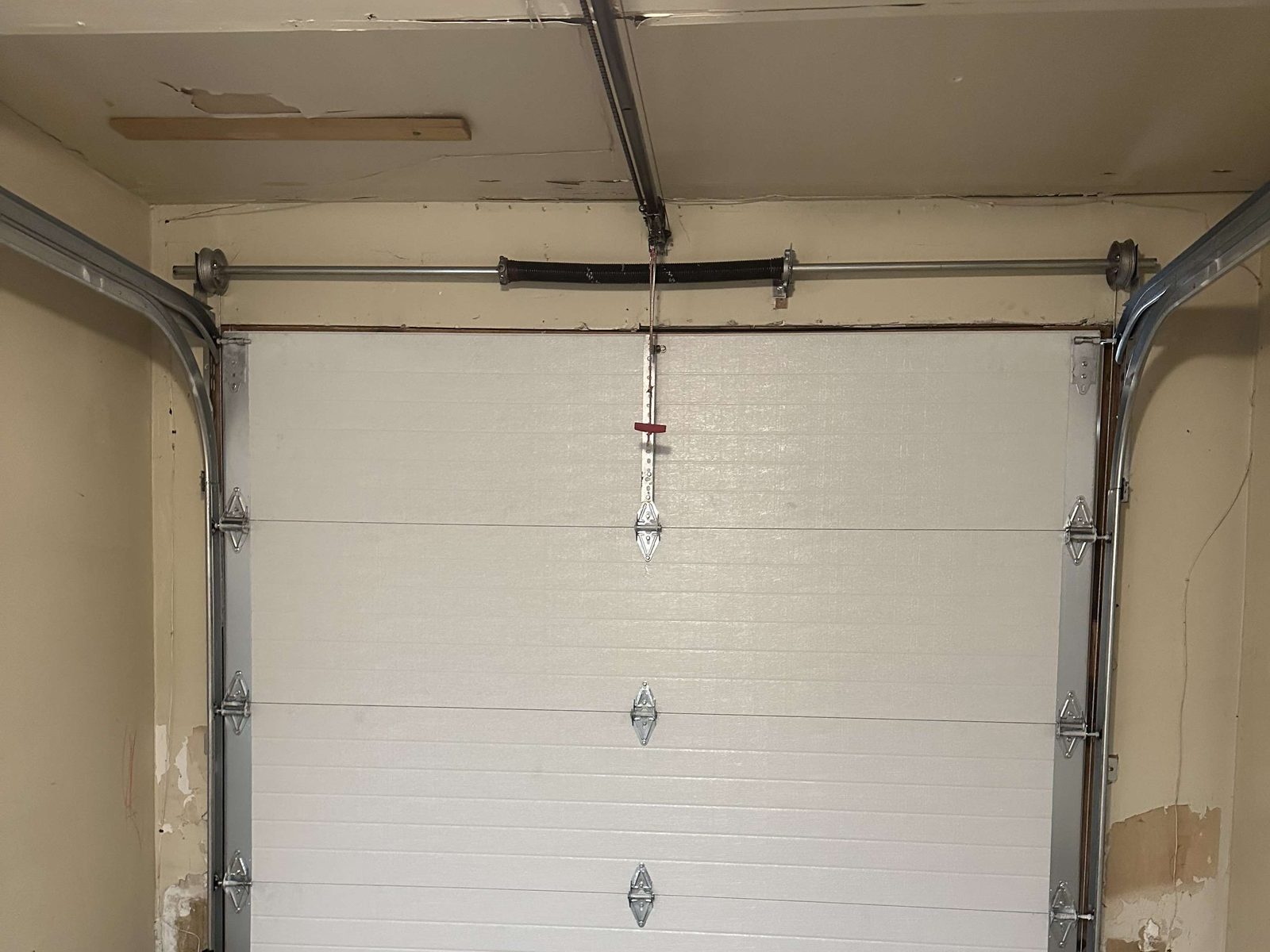 A torsion spring wound on its shaft running across the header above a closed white sectional garage door, with a cable drum at each end of the shaft and the red emergency release cord hanging below it