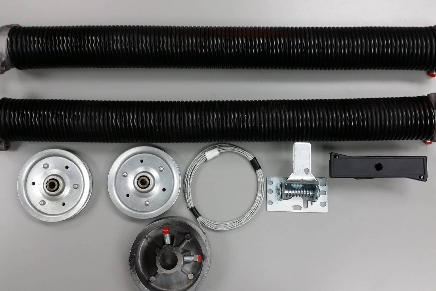 A residential garage door counterbalance kit laid out — two black torsion springs, two cable drums, a centre bearing plate, a coil of lift cable, an end bearing plate and a hinge