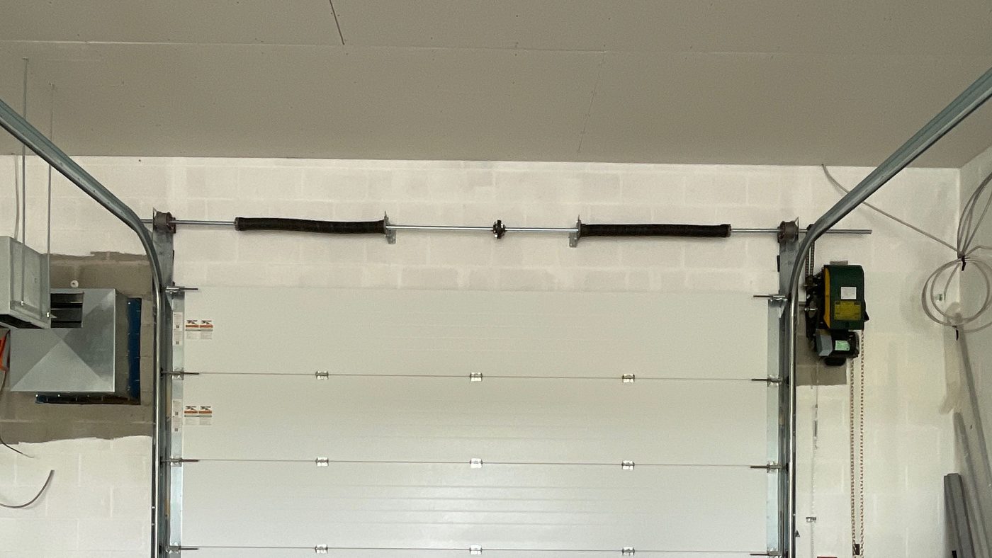 A torsion shaft mounted on the wall above a closed white sectional door, carrying two springs either side of a centre bracket, with a cable drum at each end of the shaft and a jackshaft operator on the right-hand wall