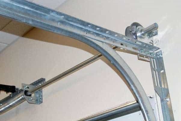 The curved section of a garage door track meeting the horizontal rail, with the cable drum and end bearing on the torsion shaft above it