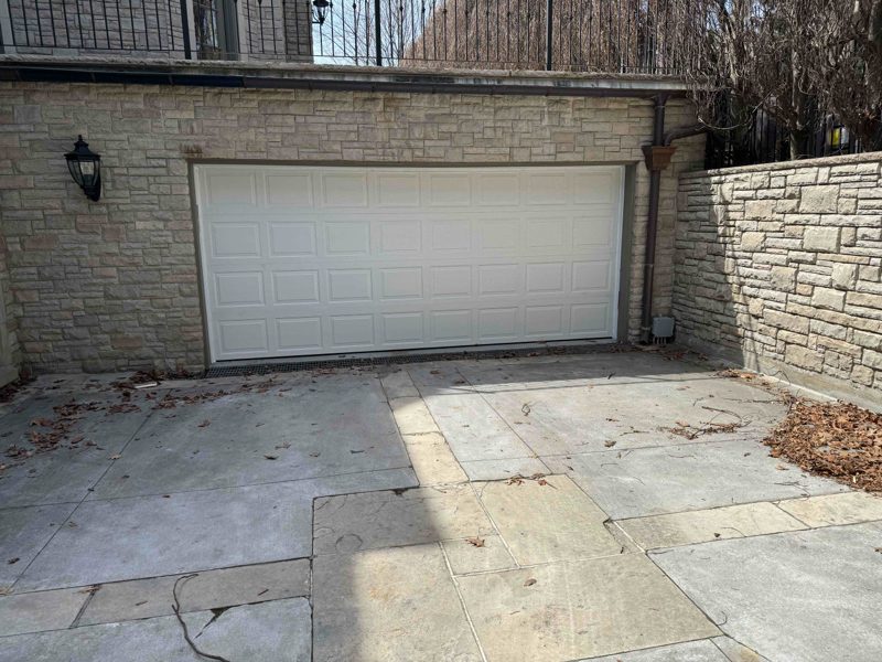 A white raised-panel double garage door fitted in a stone-clad house elevation, seen across the flagstone driveway in front of it