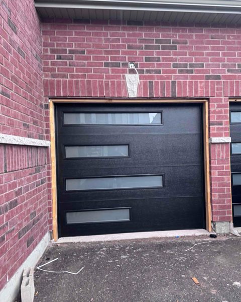 A black garage door with four frosted glazed panel slots, seen from the driveway of a new-build house, with the neighbouring door just visible beyond the brick pier