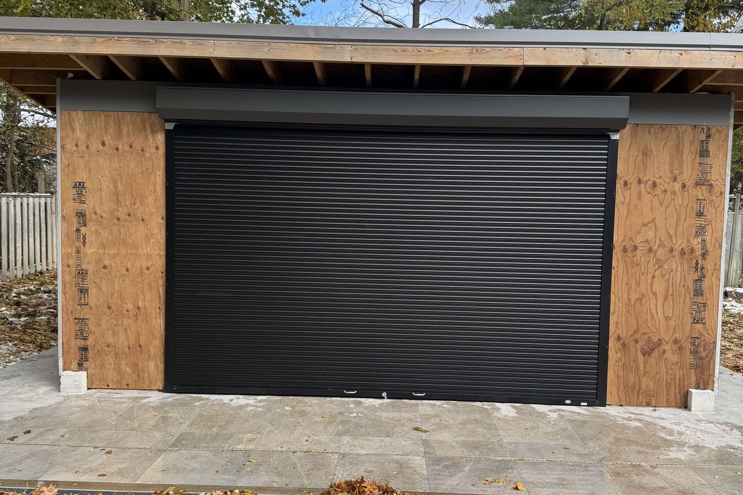 A black aluminum rolling shutter closed across the full opening of an unfinished timber-framed garden building, with the head box running the width of the opening above it