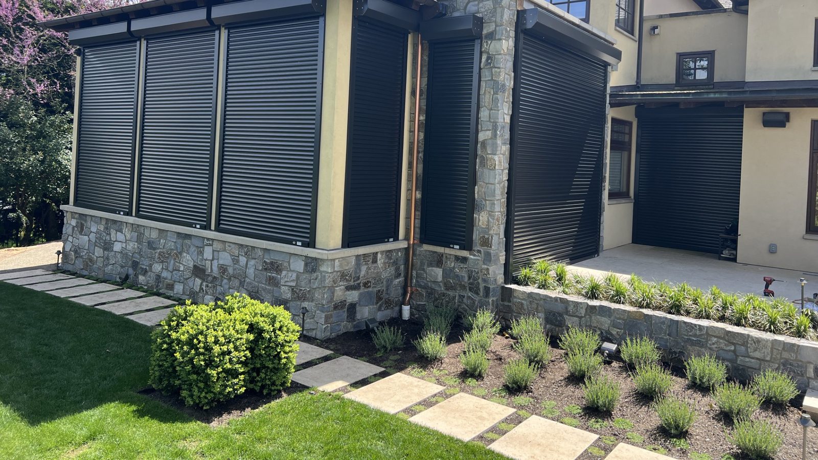Black aluminum rolling shutters closed over every window along the side of a stone and stucco house, with a matching shutter down over the garage opening beyond
