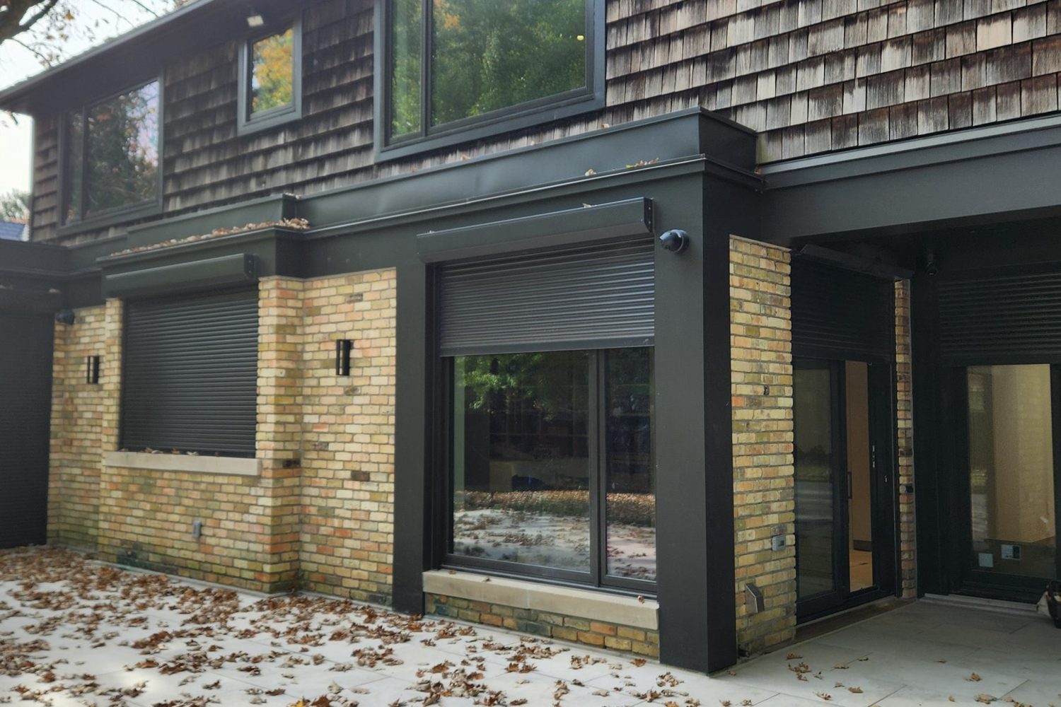 The rear elevation of a brick house with dark grey rolling shutters in built-in boxes above the windows, one shutter fully down over a window and another part-way down over a sliding door
