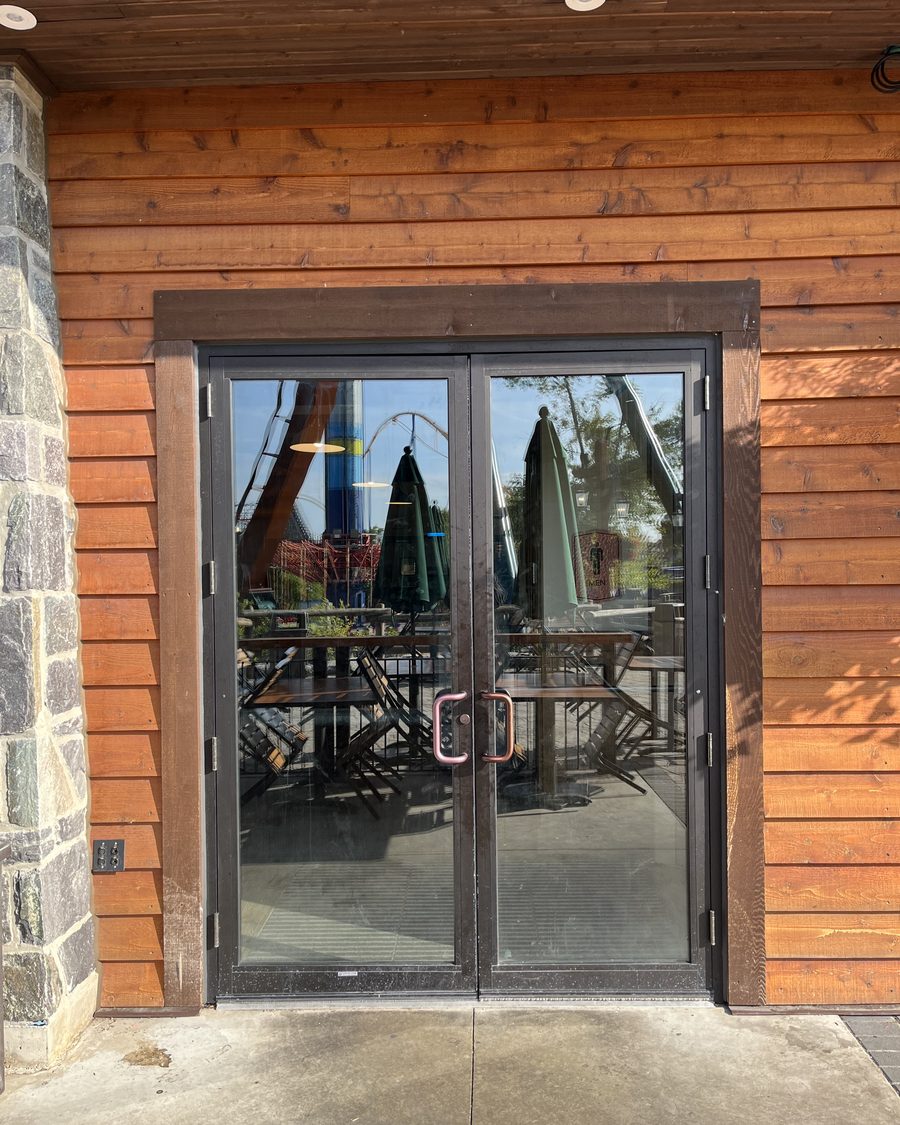 A pair of dark bronze aluminum-framed glass doors with copper-toned pull handles, set in a wood-clad wall beside a stone pillar, with patio tables, folded umbrellas and a roller coaster reflected in the glass
