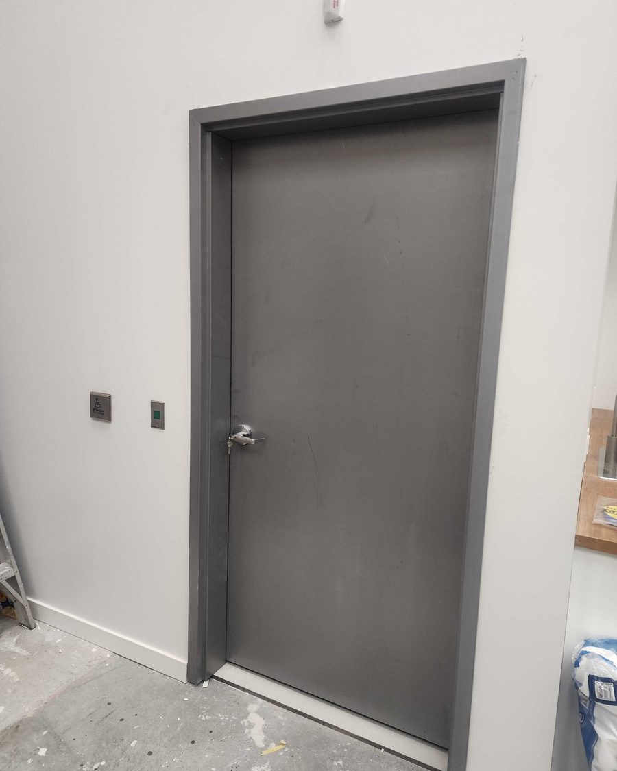 A grey steel door in a grey steel frame in a room still under construction, with a stainless push plate marked with the wheelchair symbol and a second square button with a green light mounted on the wall beside the latch side