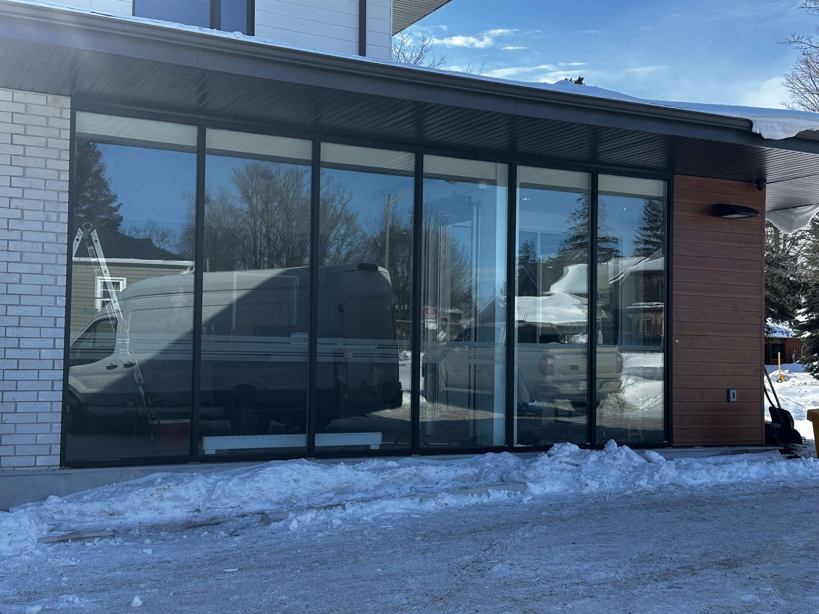 A single-storey run of six tall glass lites in black aluminum framing, set between a white brick wall and wood-look cladding under a dark soffit, with snow banked along the base and a work van and a pickup truck reflected in the glass