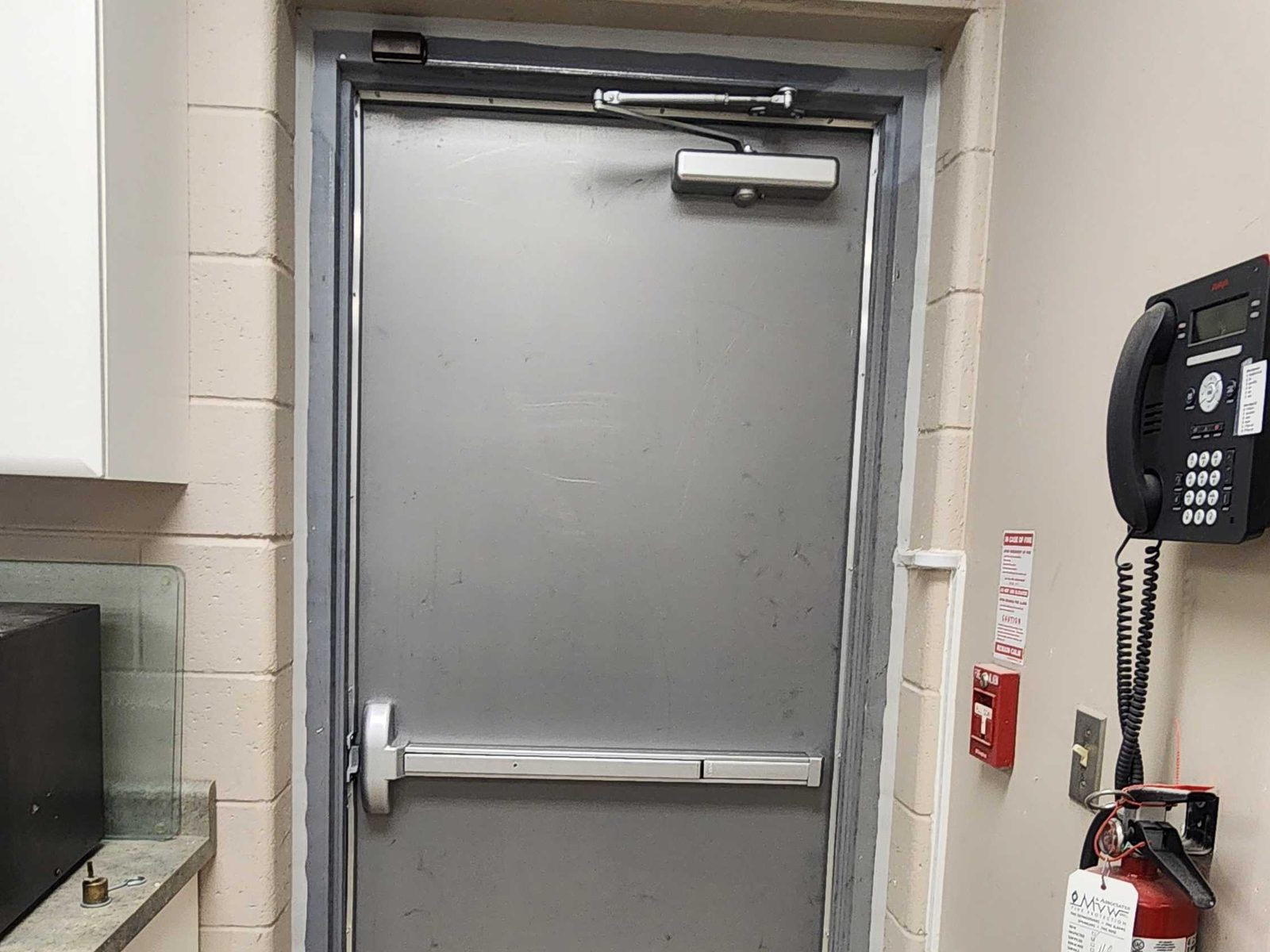 A grey steel door in a cream block-wall opening, with a surface-mounted door closer and its arm across the top of the door, a horizontal exit device across the middle, and a fire alarm pull station and wall phone beside it
