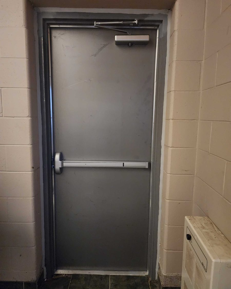 A grey steel door in a cream block-wall opening, with a surface-mounted closer at the top of the door and a horizontal exit device across its middle