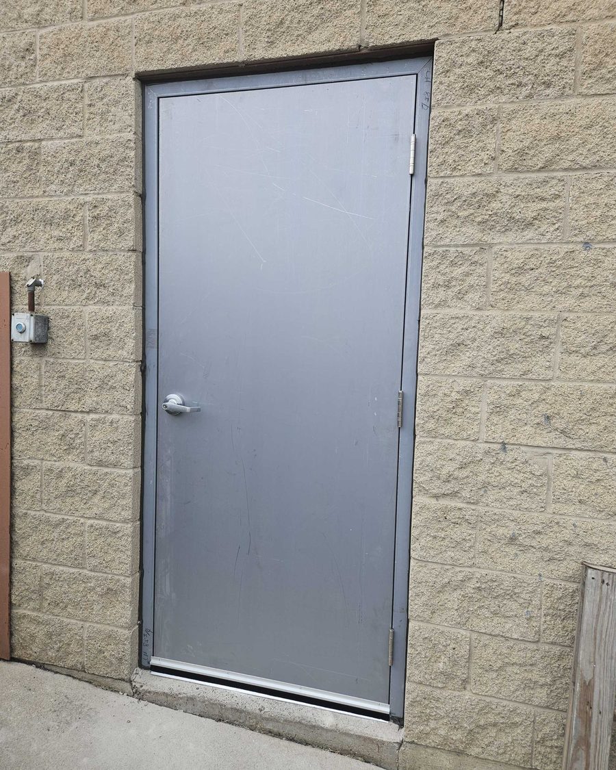 A single flush grey steel door with a lever lockset, three hinges and an aluminum threshold, in a steel frame set into a split-face block wall