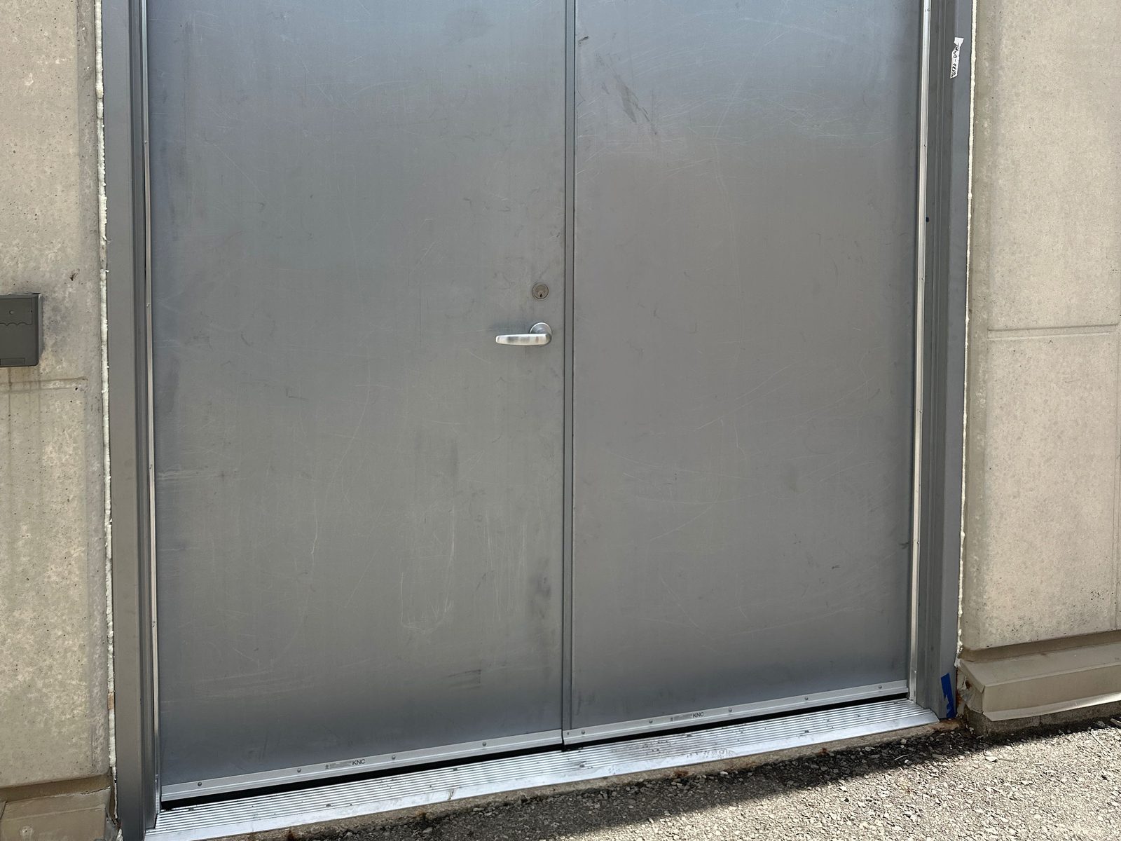 A pair of flush grey steel doors in a steel frame set into a precast concrete wall, with a lever and lock cylinder on the left leaf only, no hardware on the right leaf, a door bottom on each leaf and an aluminum threshold across the full width of the opening