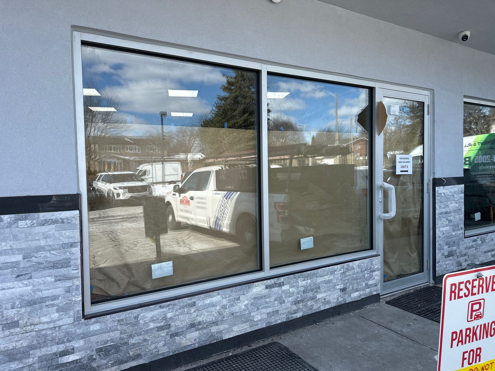 Two large storefront glass bays and a glass door in clear aluminum framing above a grey stone base, with small stickers still on the new glass, brown paper covering the floor inside, and a white pickup truck and parked cars reflected in the panes