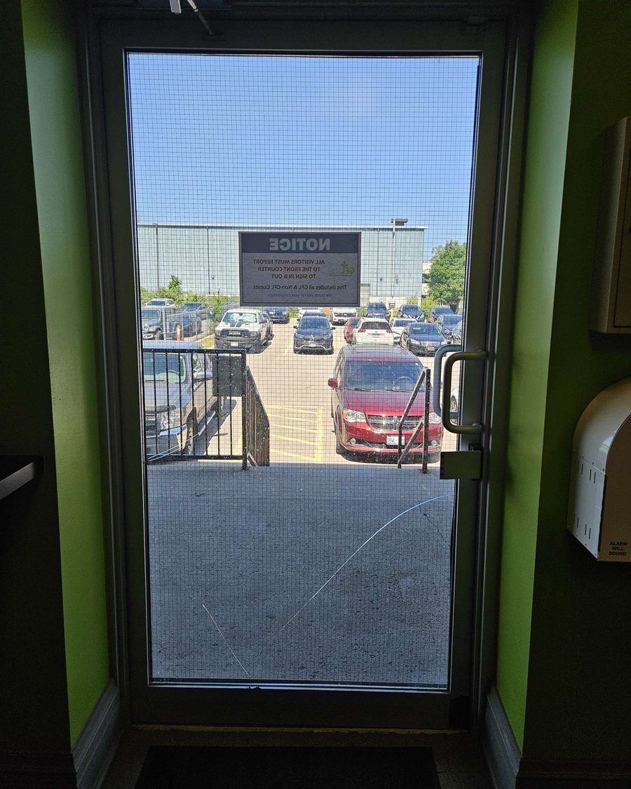 Looking out through an aluminum door with a wired-glass lite, a long crack running across its lower half, toward a parking lot and an industrial building, with a notice sign posted on the glass