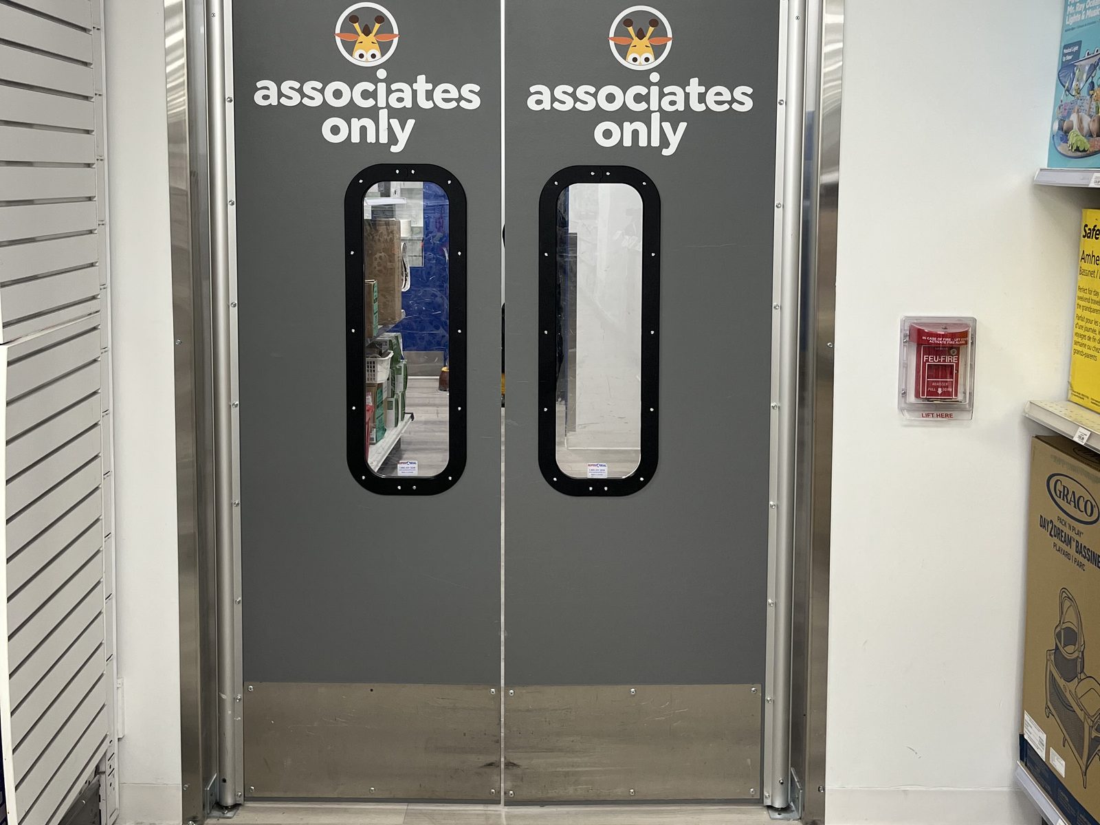 A pair of grey double-acting traffic doors marked "associates only" on a retail stockroom opening, with rounded vision windows at eye level, brushed stainless kick plates across the bottom and stainless jamb guards down both sides of the frame