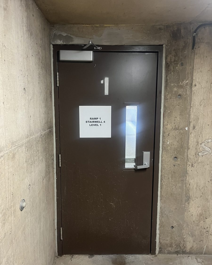 A dark brown steel stair door numbered 1 in a concrete parking structure, with a surface-mounted closer on the frame head, a lever handle, a narrow vision lite and a posted level sign
