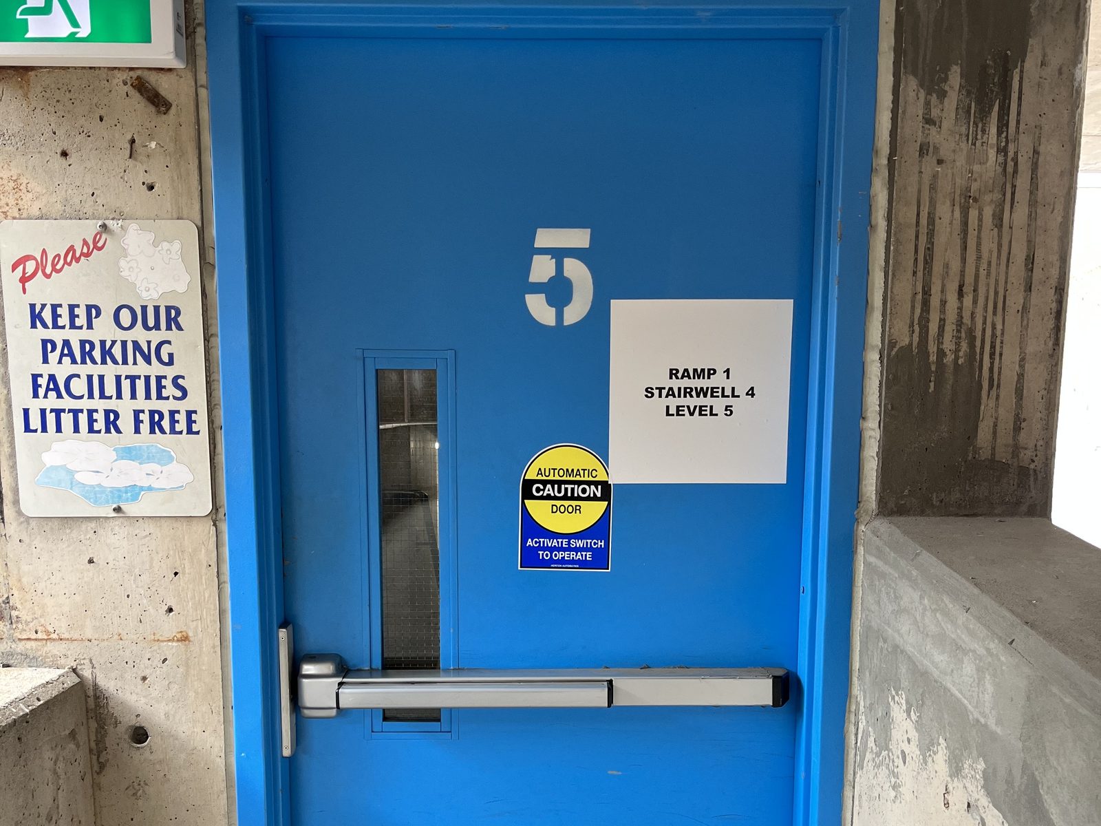 A blue-painted steel stair door numbered 5 in a concrete parking structure, with a horizontal exit device, a narrow vision lite, a posted "Ramp 1, Stairwell 4, Level 5" sign and a yellow automatic-door caution sticker on its face, and the corner of a green exit sign above