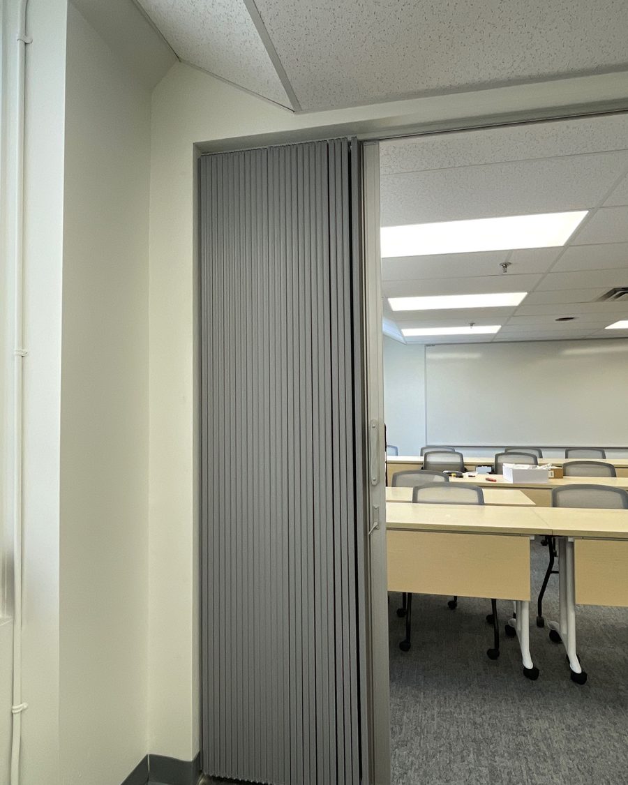A grey folding accordion partition stacked open against the jamb of a wide opening, with a training room of tables and chairs visible beyond it