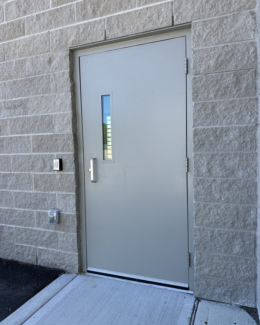 A grey-painted steel door with a narrow vision lite and a pull handle, hung in a steel frame in a split-face block wall, with a keypad and a wall light beside it and a sweep along the bottom of the door