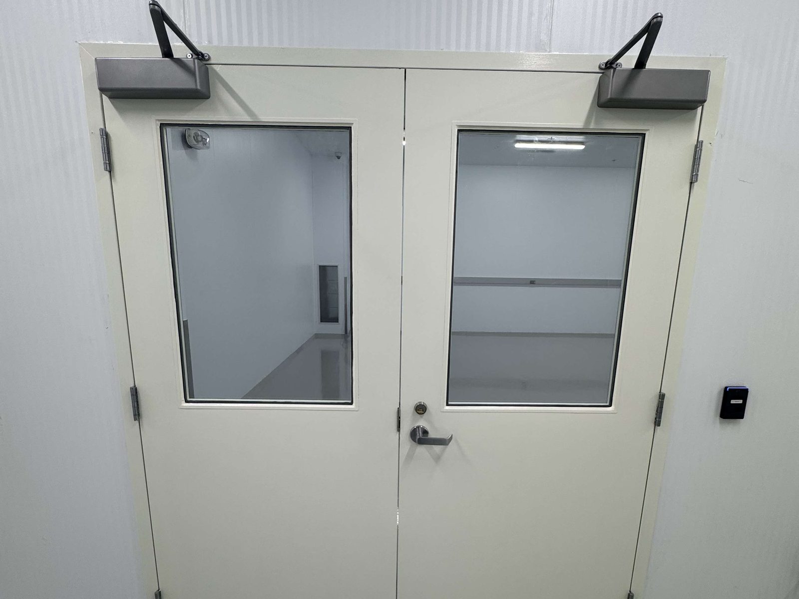 A pair of cream-painted hollow metal doors in a steel frame, each leaf with a large vision lite and a surface-mounted closer along its top, a lever handle on the right leaf and a card reader on the white panelled wall beside it