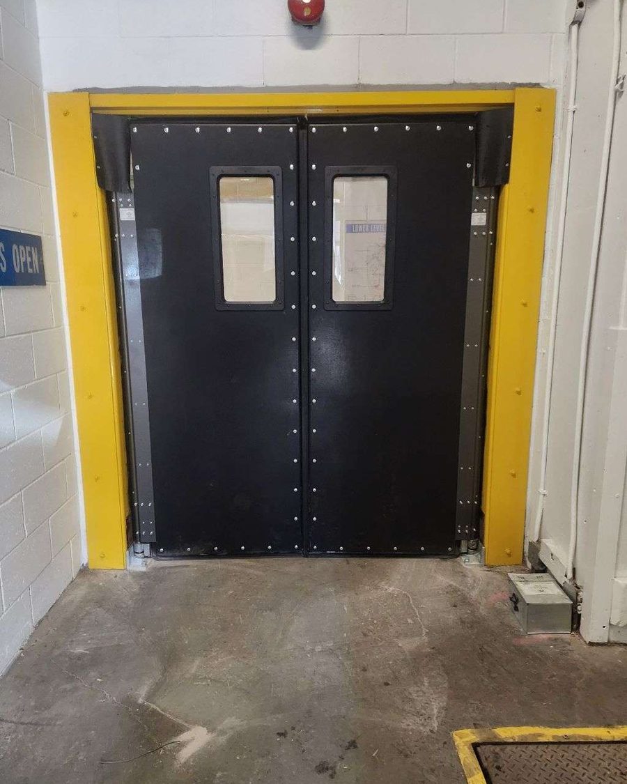 A pair of black double-acting impact doors with small rectangular vision windows, set in a yellow-painted steel frame in a concrete-block warehouse wall above a bare concrete floor