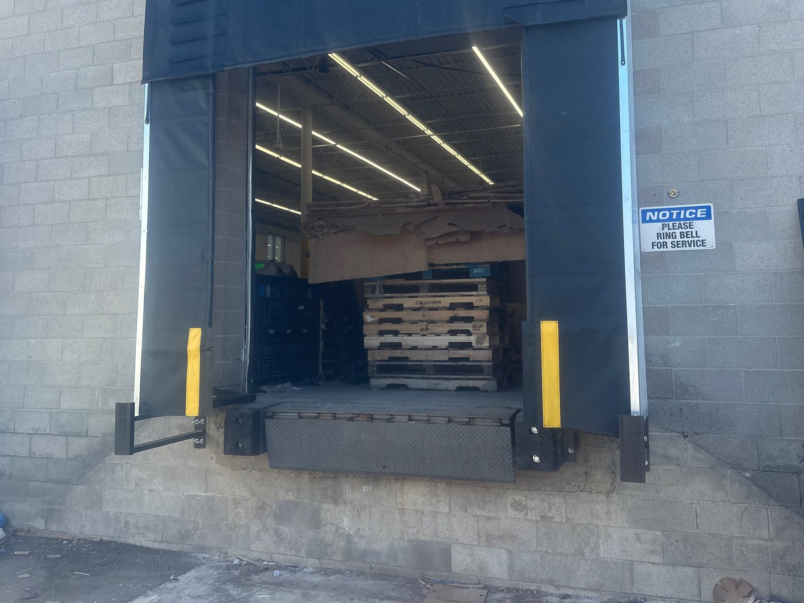 A loading dock position in a grey block wall, its floor raised well above the concrete apron, fitted with a black fabric dock shelter with yellow-striped side curtains, black rubber bumpers either side and a diamond-plate edge-of-dock leveler lip hanging on the dock face, with stacked pallets inside and a "Please ring bell for service" notice on the wall