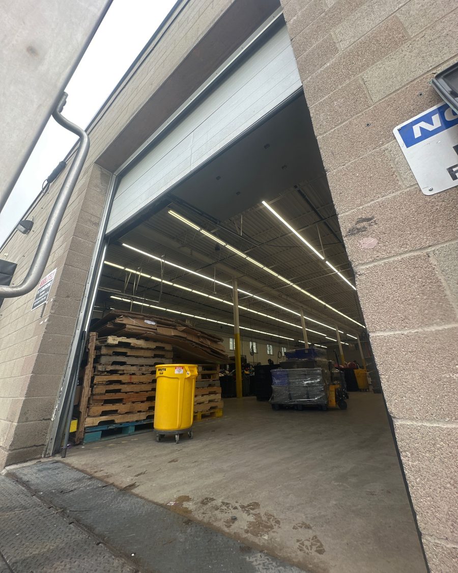An open dock door seen from the apron, its white sectional door rolled up into the head, looking into a warehouse with stacked wooden pallets, a yellow waste bin and wrapped loads under strip lighting, with diamond-plate steel at the dock edge in the foreground