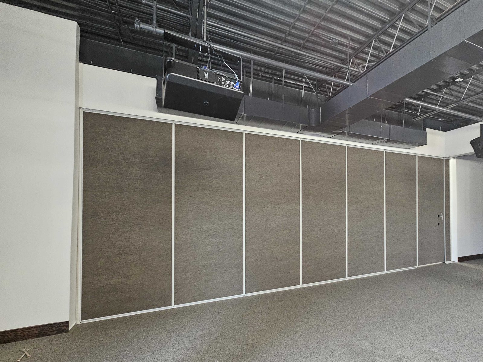 A closed operable wall of dark fabric-faced panels in light edge trim across a carpeted hall, with a pass door and lever handle in the right-hand panel, a solid header wall above the wall, and an exposed steel roof deck with a hanging projector overhead