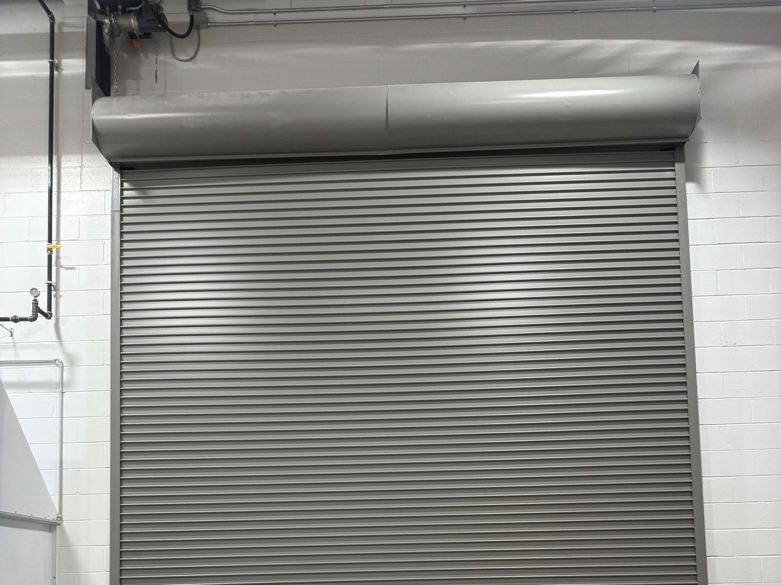 The top half of a grey rolling steel door inside a white block-walled building: a rounded steel hood across the head of the opening, the slatted curtain below it, a guide down each side, and drive equipment mounted on the wall above the left end of the hood