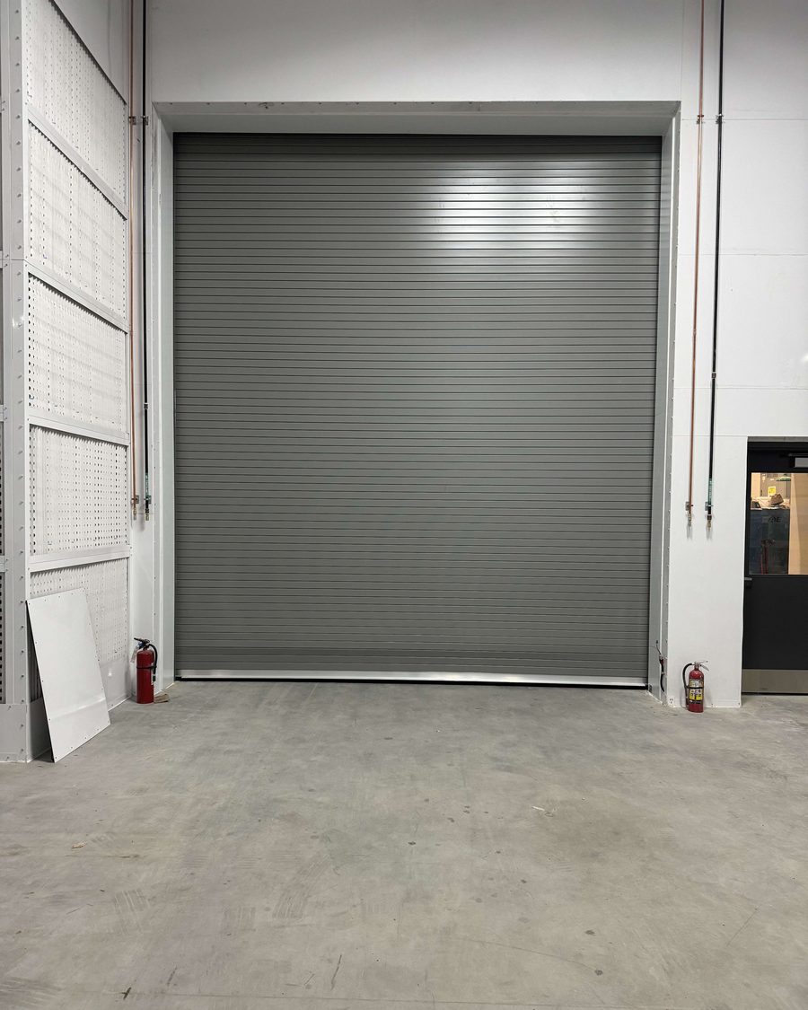 A grey rolling steel door closed in a tall opening inside a warehouse, with a red fire extinguisher on the floor at each side of the opening and a concrete floor in front