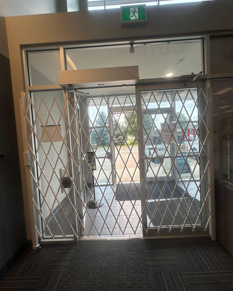 A white diamond-pattern folding security gate drawn across a glazed shop entrance, with the vestibule doors, a green exit sign above and a parking lot visible through it