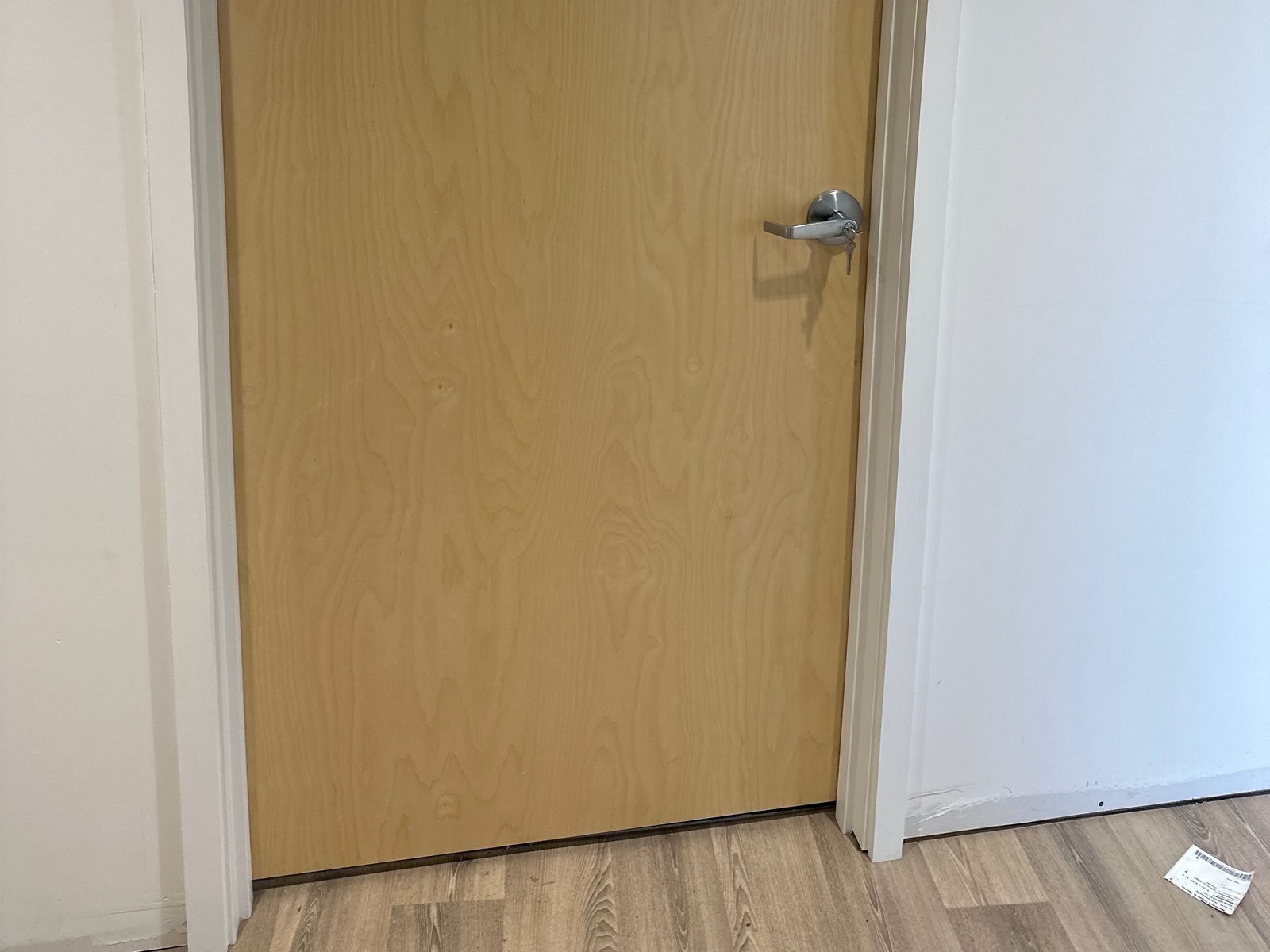 A flush light wood-veneer office door, closed in a white-trimmed frame, with a satin chrome lever and cylinder, and a thin dark gap between the bottom of the door and the vinyl plank floor