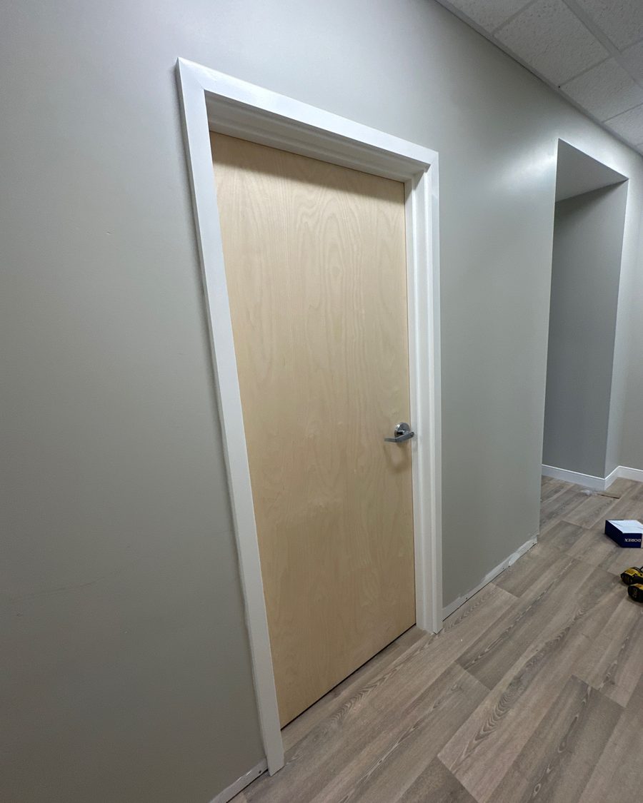 A light wood-veneer office door with a lever handle in a white-trimmed opening along a grey corridor with a dropped ceiling and vinyl plank floor