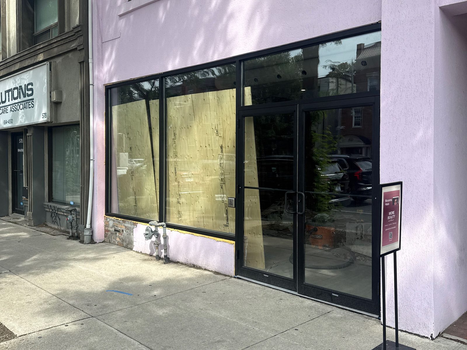 A new black aluminum storefront on a lilac-painted street-front building: two large fixed glass bays, a transom lite running above a pair of glass entrance doors, and plywood sheets leaning against the inside of the glass while the unit is fitted out
